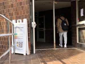 A student walks into Wilson Commons, passing a sign detailing the University's former tiered masking guideline system. The sign is folded up and placed between a railing and a brick wall, looking somewhat discarded.