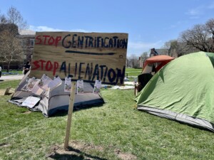 A wooden sign that reads "Stop Gentrification. Stop Alientation" in front of some tents on Wilson Quad.